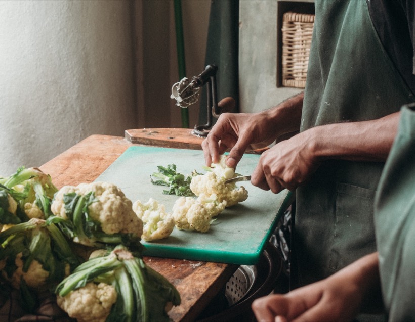 Hands trimming cauliflower on a chopping board next to a hand-cranked coconut scraper in the Lanka Yoga kitchen