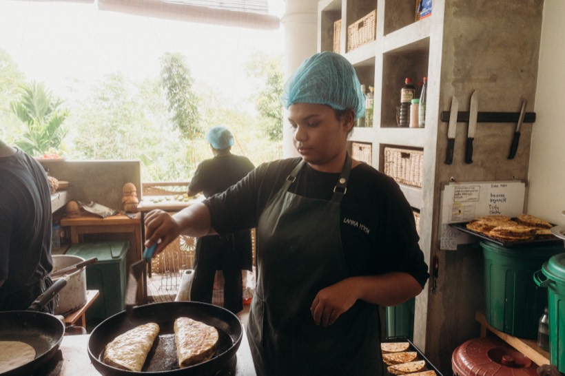 A cook in a Lanka Yoga apron turning pol roti in a pan in the open kitchen, with a tray of cooked roti stacked beside her