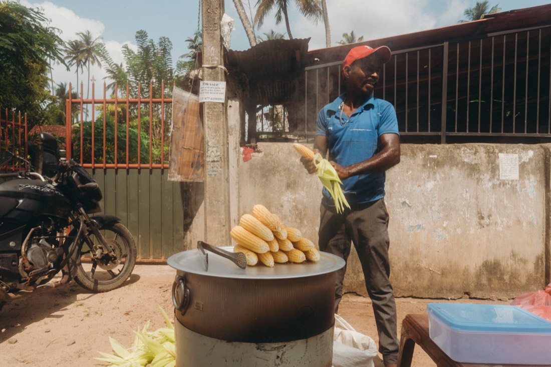 A roadside vendor in Sri Lanka peeling corn beside a steaming pot of boiled cobs, with a motorbike and palm trees behind him