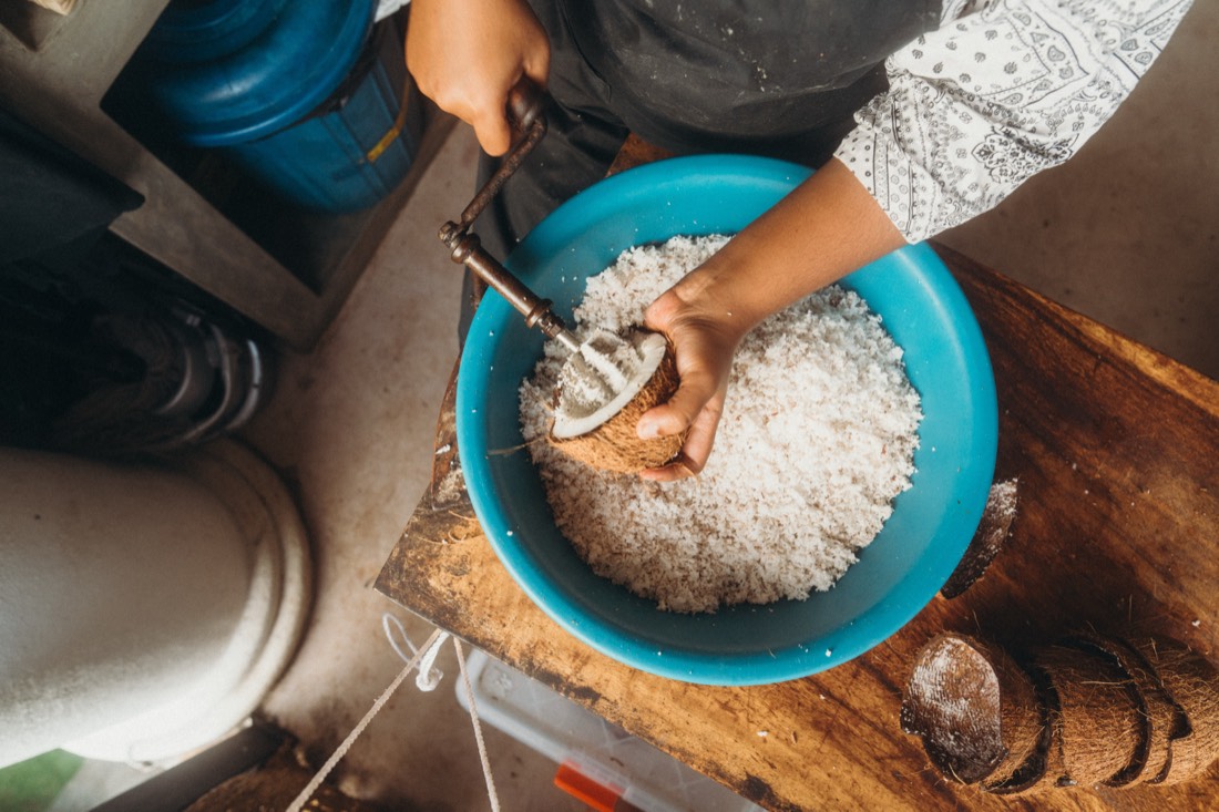 A cook scraping the flesh from half a fresh coconut on a hand-cranked grater into a bowl of shredded coconut, ready for pol sambol