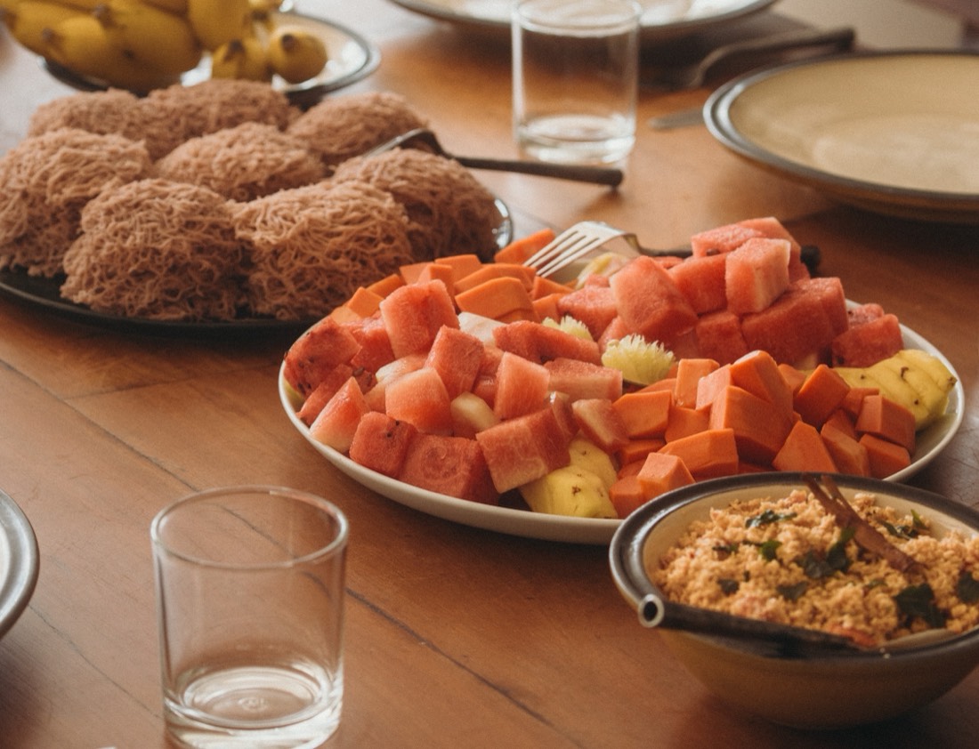 Breakfast on a wooden table in Sri Lanka: a plate of steamed red rice string hoppers beside cut watermelon, papaya and pineapple, with a bowl of coconut sambol