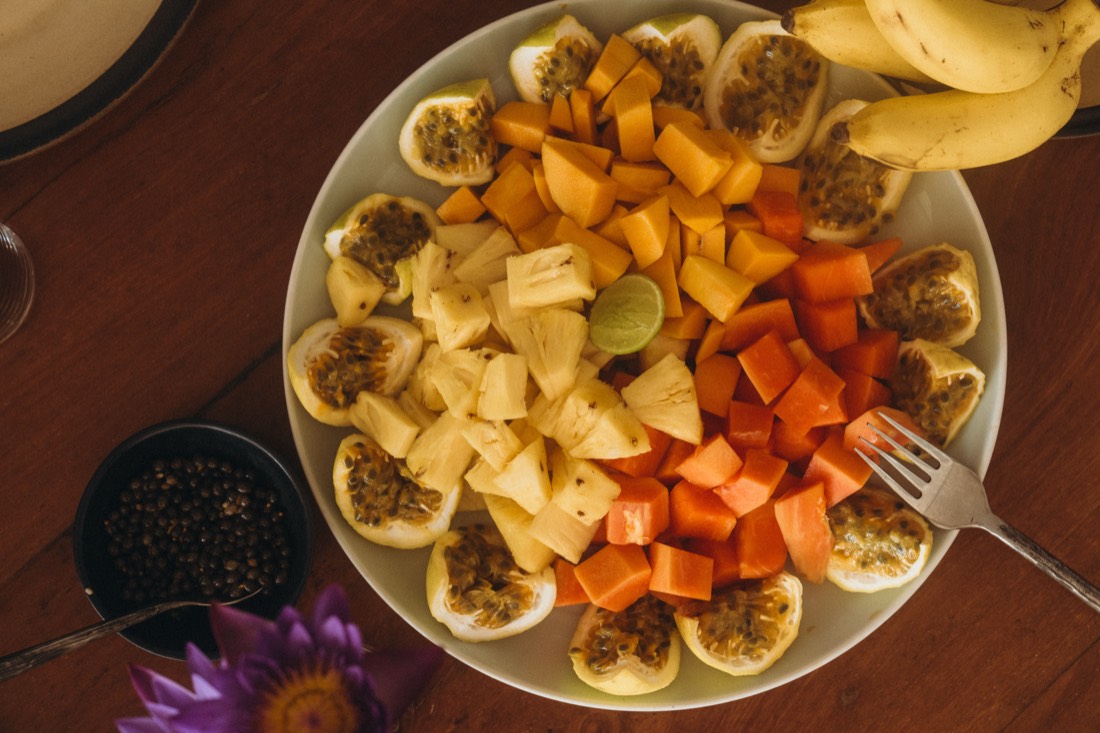 A platter of cut mango, pineapple and papaya ringed with halved passionfruit and a wedge of lime, on a wooden table at Lanka Yoga